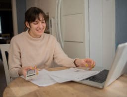 Egy fiatal nő kalkulál az előtte lévő laptopon, számológéppel, és papírokat néz.