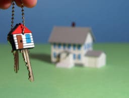 Előtérben egyy ház alakú kulcstartót tart valaki az újján lakáskucsokkal, háttérben egy ház makett.