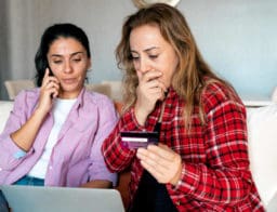 két nő ül a kanapén, laptop előtt, banki csalás áldozatává váltak, bejelentést tesznek telefonon