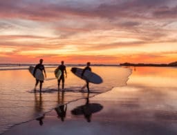balesetbiztosítás és utasbiztosítás közti különbség: három ember a vízparton, naplementében, szörfdeszkával a kezében, nyaralás során