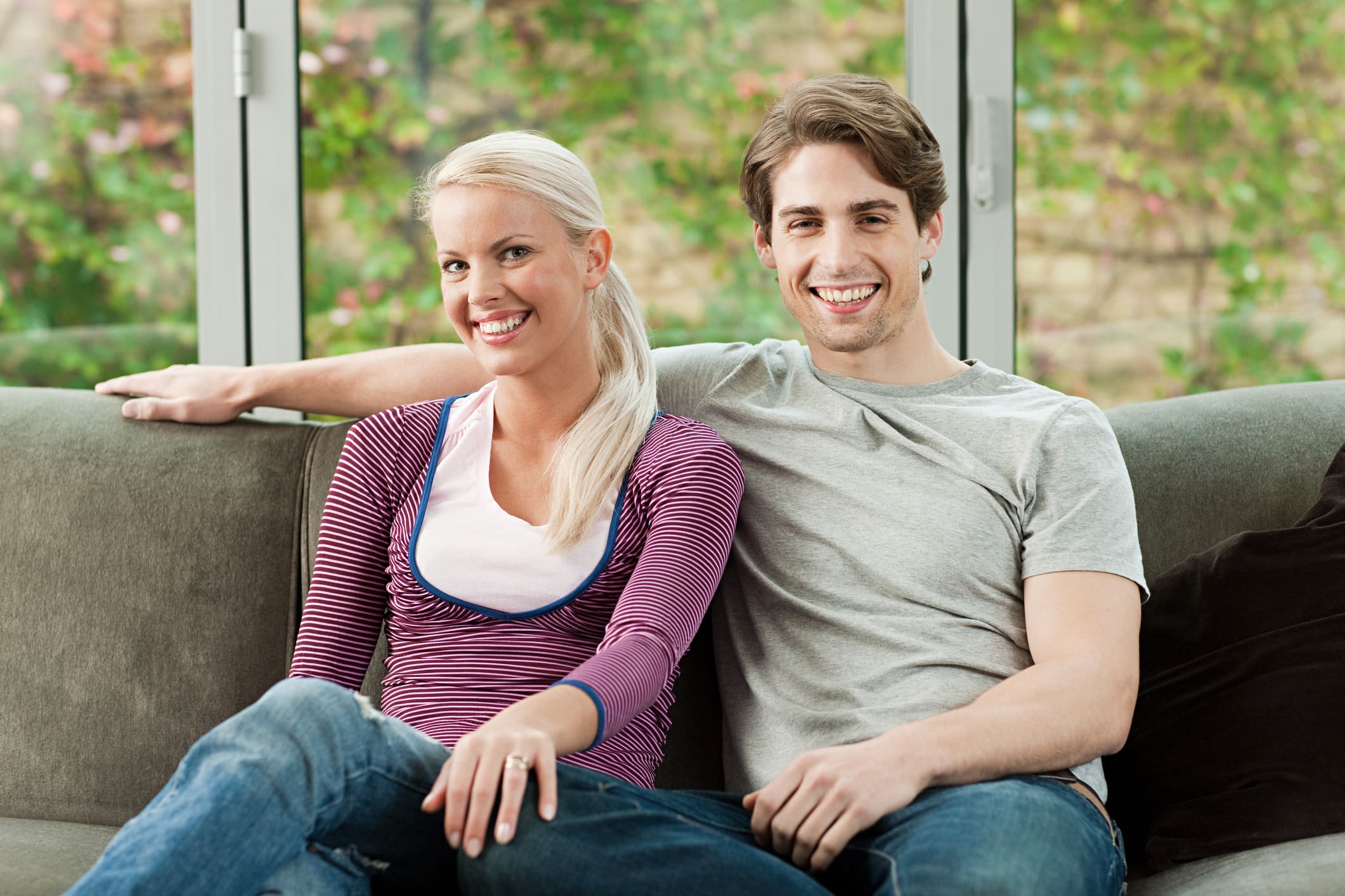 Fiatal házaspár ül egy kanapén a nappaliban, mögöttük üvegajtó, természet.