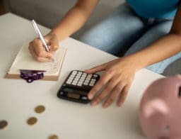 Íróasztalon malacpersely, pénzérmék, számológép, jegyzetfüzet, egy nő tollal jegyzetel, közben számol