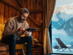 középkorú férfi faházban, odakint havas hegyek, kezében laptop, tél van, a nyaralására készül