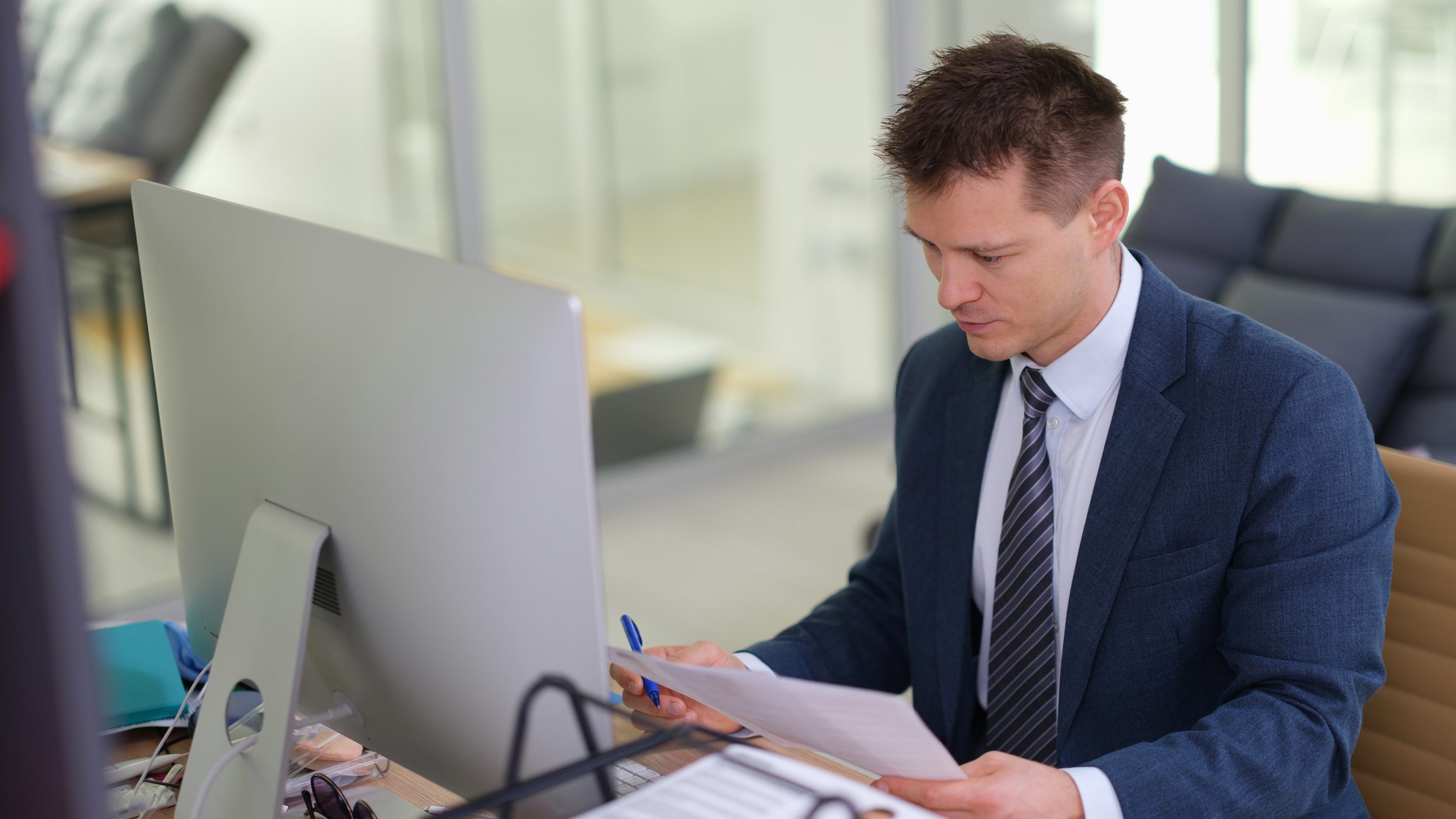 Egy öltönyös férfi egy irodában a számítógépnél ül a munkaasztalánál és egy papírlapot tanulmányoz.